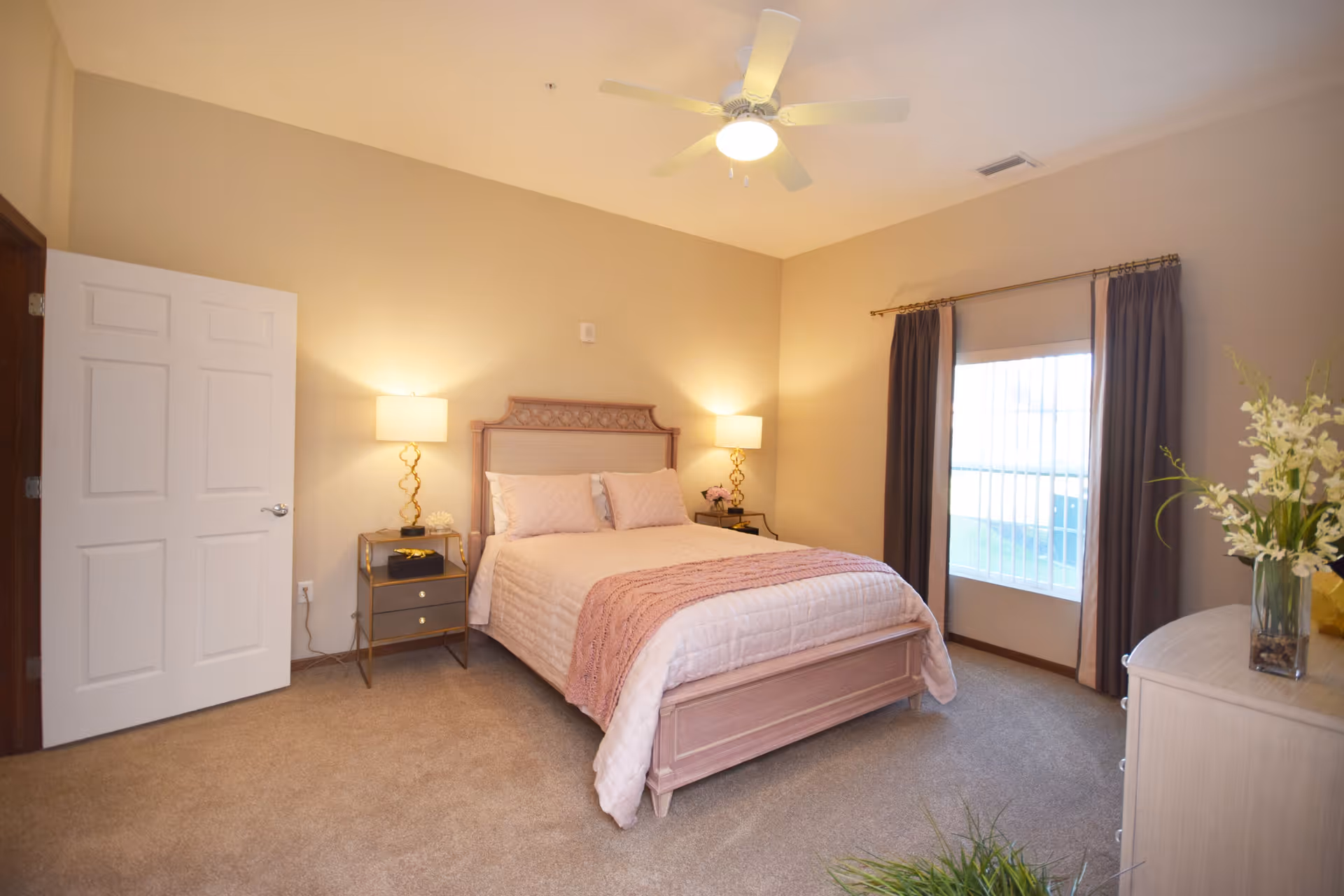 A cozy bedroom with a queen-sized bed featuring a light pink quilt and matching pillows. Two bedside tables with lamps flank the bed. There is a window with dark curtains on the right side, and a ceiling fan with a light is mounted on the ceiling. A dresser with a vase of flowers is visible on the right side of the room. The walls are painted beige and the floor is carpeted.