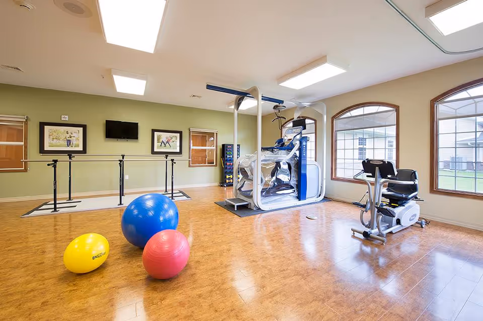 A bright exercise room with wooden flooring featuring three exercise balls in yellow, blue, and red on the floor. The room has large windows with arched tops letting in natural light. There is exercise equipment including a recumbent bike and a large machine with support bars. The walls are light green and beige with framed pictures and a wall-mounted TV.