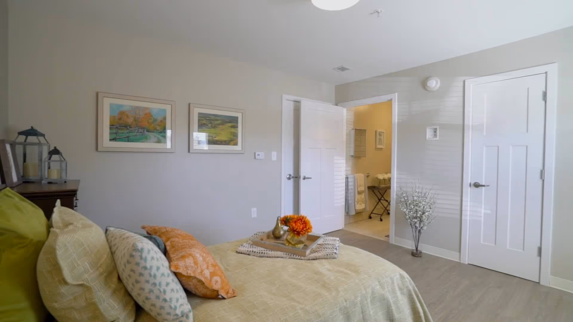 A bedroom with a neatly made bed featuring multiple pillows and a tray with a flower arrangement on top. The room has light-colored walls with two framed pictures hanging. There are two white doors, one open leading to a bathroom area with towels and a small table, and the other closed. A decorative plant is placed near the closed door.