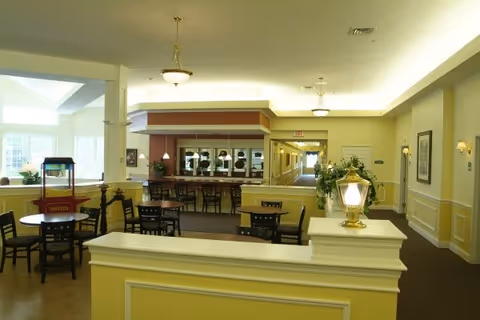 Interior view of a senior living facility common area with multiple tables and chairs arranged for dining or socializing. There is a popcorn machine on one table, decorative lighting fixtures on the ceiling, and a hallway leading further into the building. The walls are painted yellow and white with framed artwork and plants placed around.