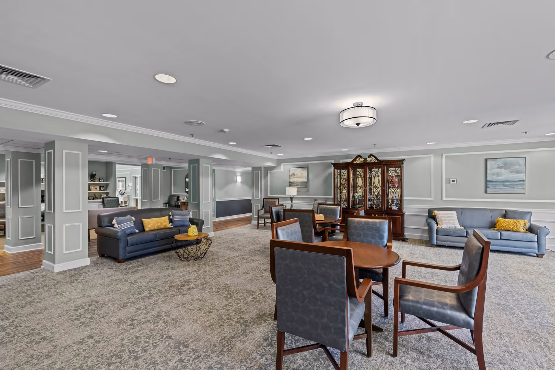 A spacious senior living common area with multiple seating options including two blue sofas with yellow and patterned pillows, a round wooden table surrounded by six cushioned chairs, and a wooden china cabinet against the wall. The room has light gray walls with white trim, carpeted flooring, and ceiling lights.