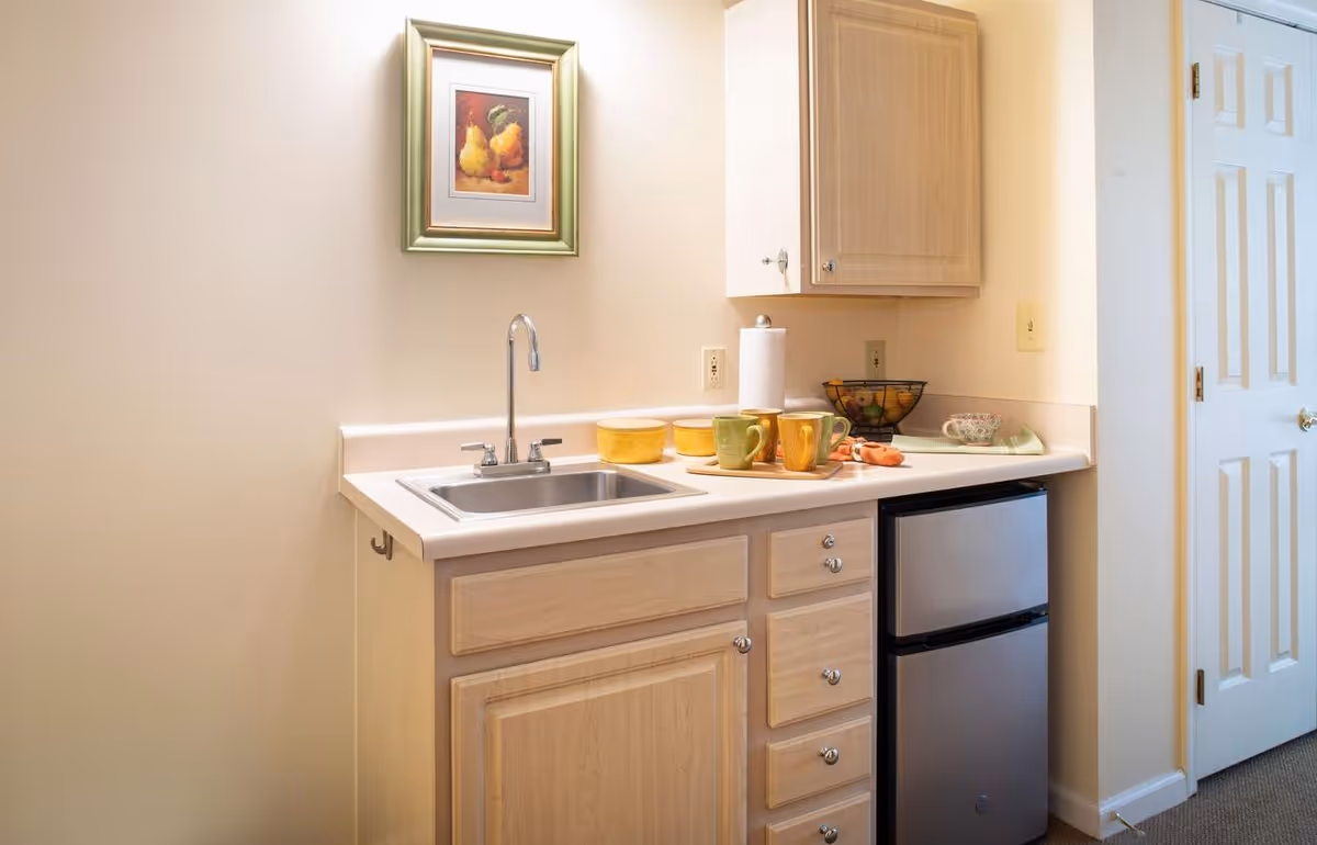 Small kitchenette with a sink, light wood cabinets, countertop mugs and fruit bowl, and a stainless steel mini refrigerator.
