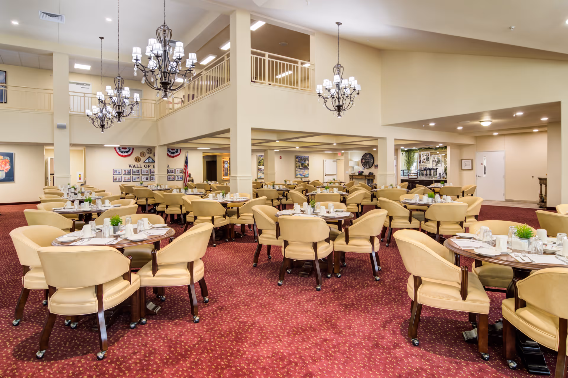 Spacious dining room with multiple round tables set with white dishes, cups, and silverware. Each table is surrounded by beige upholstered chairs with wooden legs. The room features a red patterned carpet, cream-colored walls, and several elegant chandeliers hanging from the ceiling. There is a second-floor balcony overlooking the dining area and a wall display labeled 'Wall of Honor' with framed photos and an American flag.