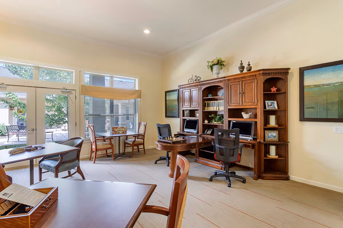 A well-lit interior room with large windows and glass doors leading outside. The room features several tables and chairs, a wooden desk with two office chairs, two laptops, bookshelves with decorative items, and framed artwork on the walls. The space appears to be a common area or office within an assisted living facility.