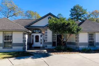 Front exterior view of a single-story senior care home building with a gabled roof, white and gray walls, multiple windows, a central entrance with a glass door, and a tree in front of the entrance.