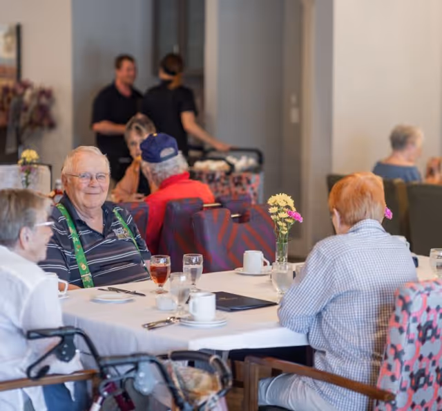 A group of elderly residents seated around a table in a dining room with drinks, flowers, and table settings.