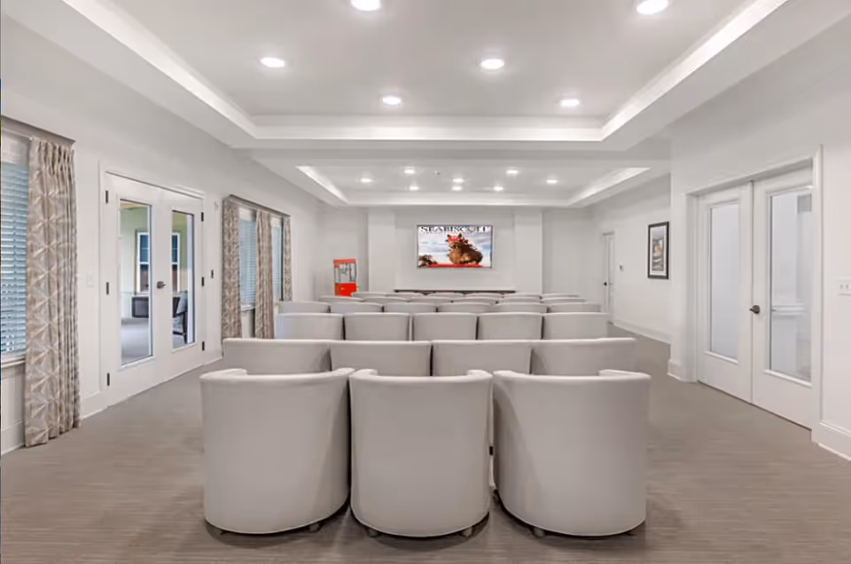 A modern media room with multiple rows of white cushioned chairs facing a large flat-screen TV mounted on the wall. The room has recessed lighting, light-colored walls, carpeted floor, and large windows with patterned curtains. There is a red popcorn machine near the TV and double glass doors on both sides of the room.