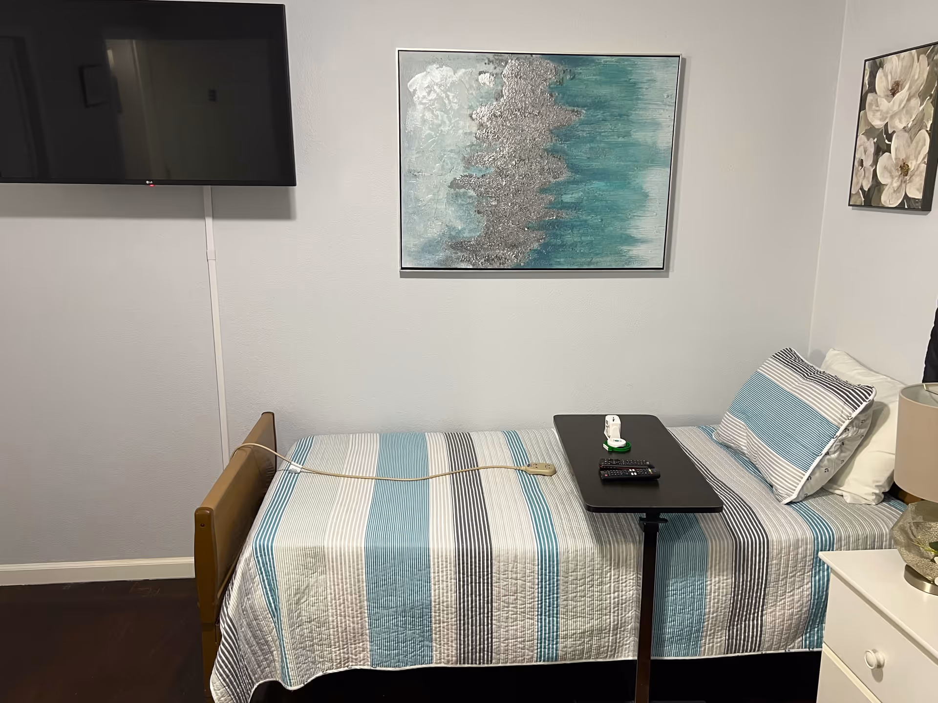 A small bedroom with a single bed covered in a striped blue, white, and gray quilt. A black overbed table holds a remote control and a small device. A flat-screen TV is mounted on the wall to the left. Two framed artworks hang on the walls, one above the bed featuring abstract blue and silver tones, and another with white flowers on the adjacent wall. A white nightstand with a lamp and decorative bowl is next to the bed.