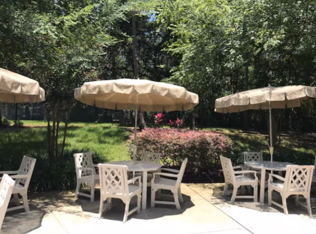 Outdoor patio area with multiple round tables, each surrounded by white chairs and shaded by beige umbrellas. The patio is adjacent to a grassy area with trees and bushes providing a green backdrop.