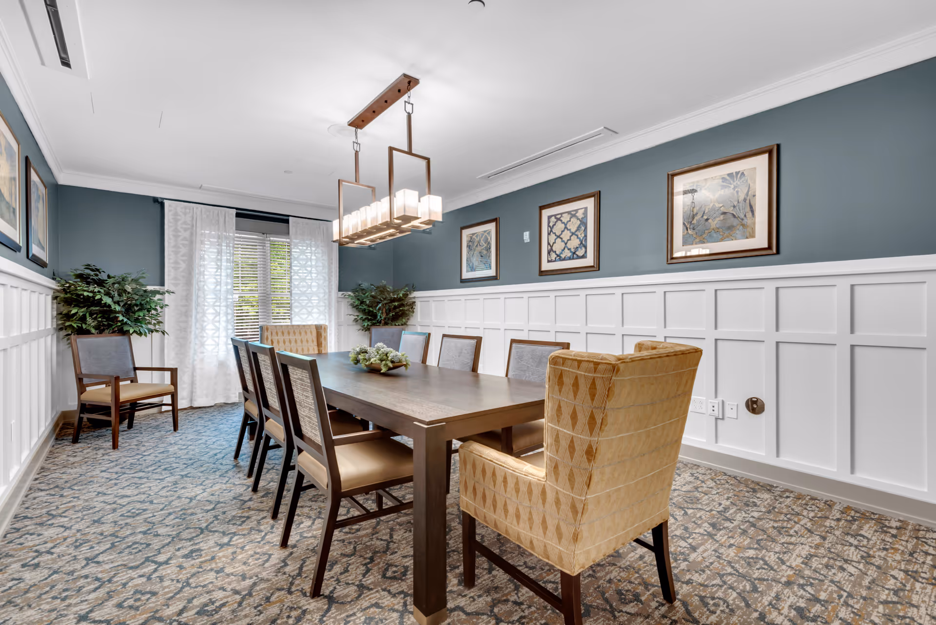 A well-lit dining room with a long wooden table surrounded by eight chairs, including two upholstered armchairs at the ends. The room features blue walls with white wainscoting, patterned carpet, framed artwork on the walls, two potted plants in the corners, and a modern rectangular chandelier hanging above the table. A window with white curtains allows natural light into the room.