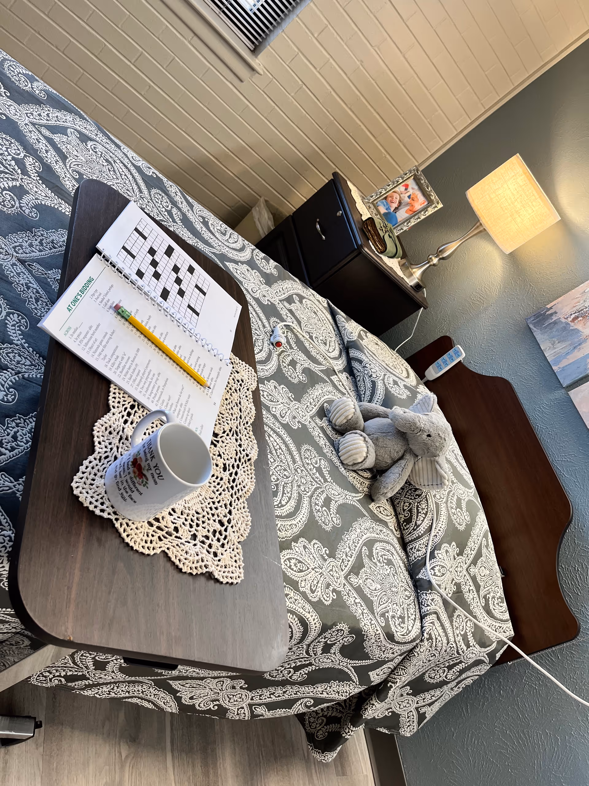 A neatly made bed with a patterned gray and white bedspread in a nursing facility room. On the bed is a small stuffed elephant toy and a wooden overbed table holding a crossword puzzle book, a pencil, and a white mug on a crocheted doily. Next to the bed is a dark wooden nightstand with a lamp, a framed photo, and some personal items. The walls are painted gray and there is a window with blinds partially visible.