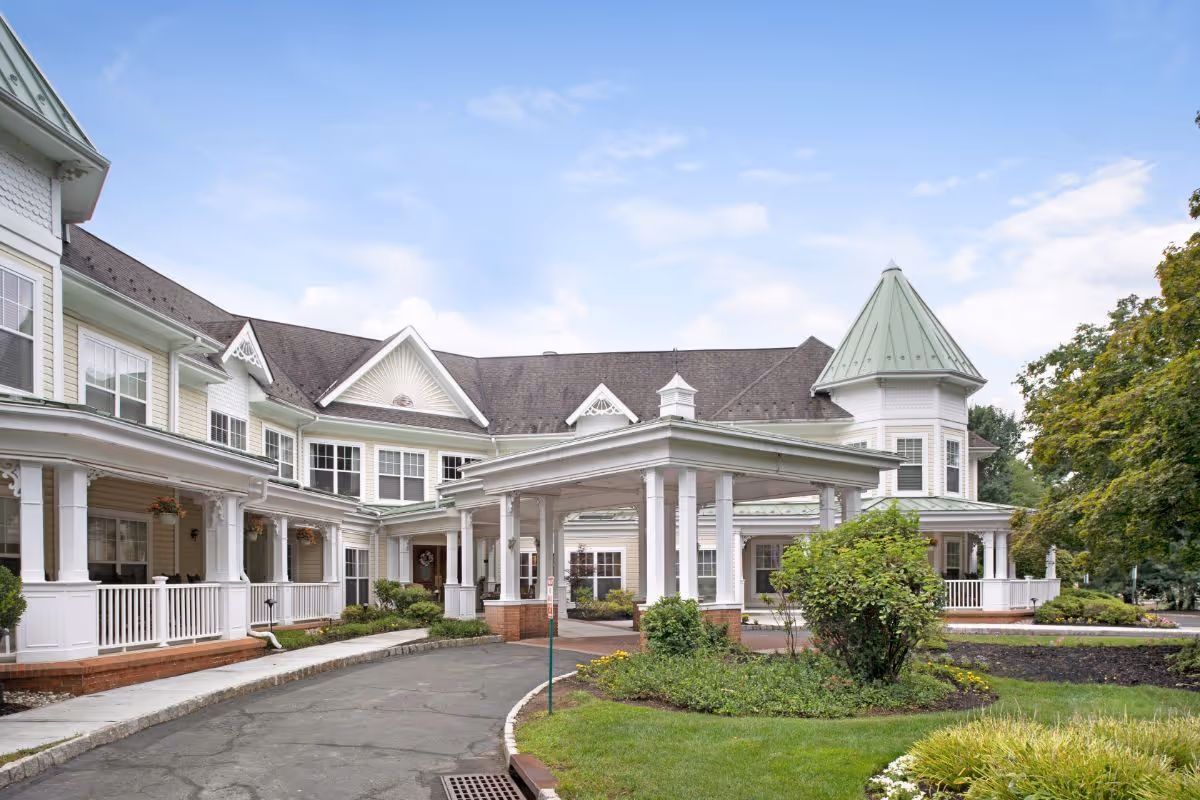 Front entrance of a large Victorian-style senior living building with a covered porte-cochere, wraparound porch, and landscaped grounds.
