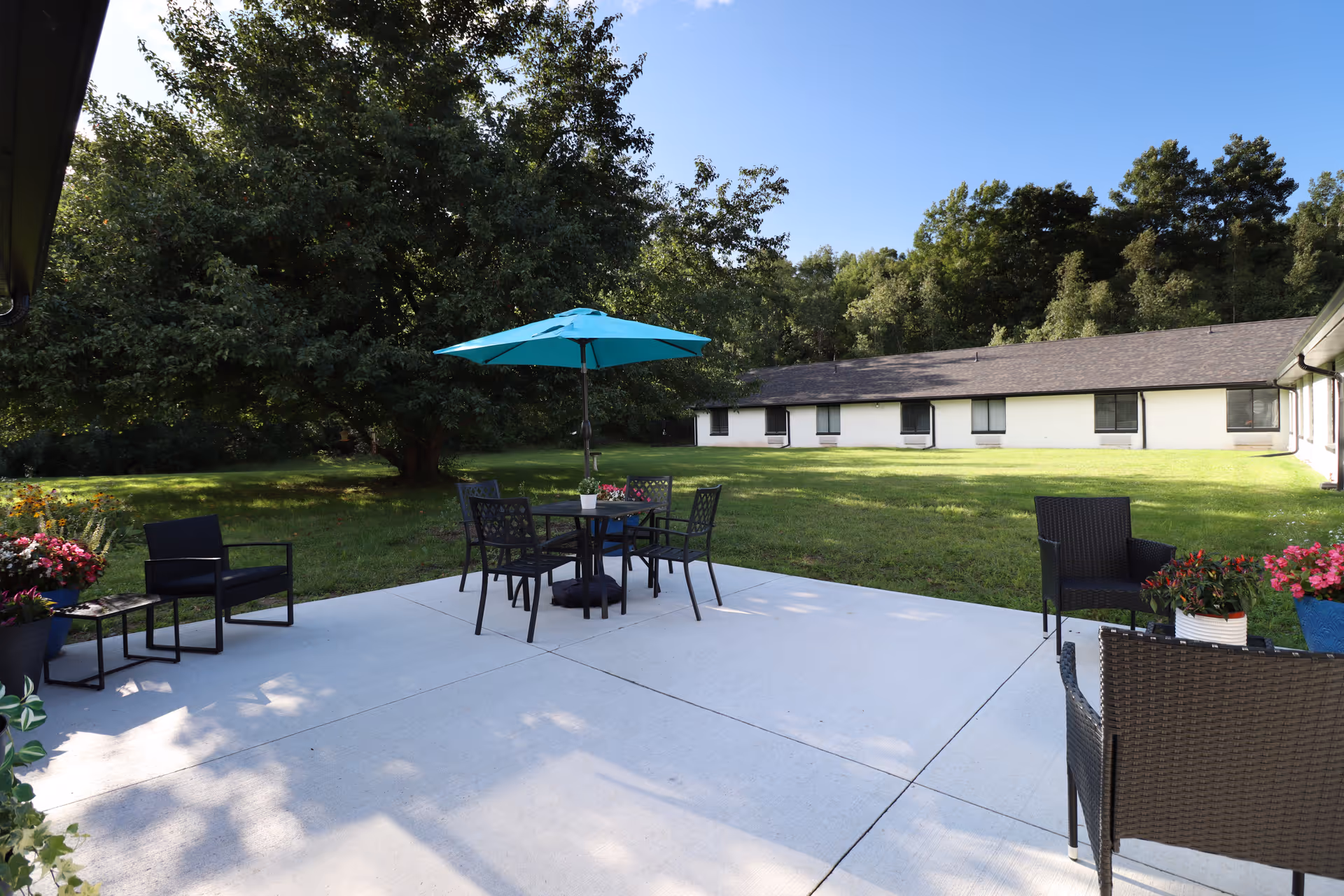 Outdoor patio with chairs and a table under a teal umbrella facing a lawn and a single-story building surrounded by trees.
