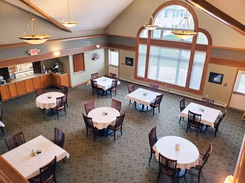 A spacious dining room with several round and square tables covered with white tablecloths, each surrounded by wooden chairs. The room features large arched windows letting in natural light, high ceilings with wooden beams, and pendant lights hanging from the ceiling. There is a small kitchenette area with cabinets and a coffee machine in one corner.