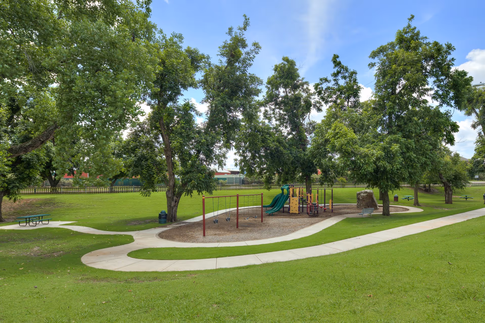 Outdoor playground area with swings, slides, and climbing structures surrounded by green grass, trees, and paved walking paths under a partly cloudy sky.