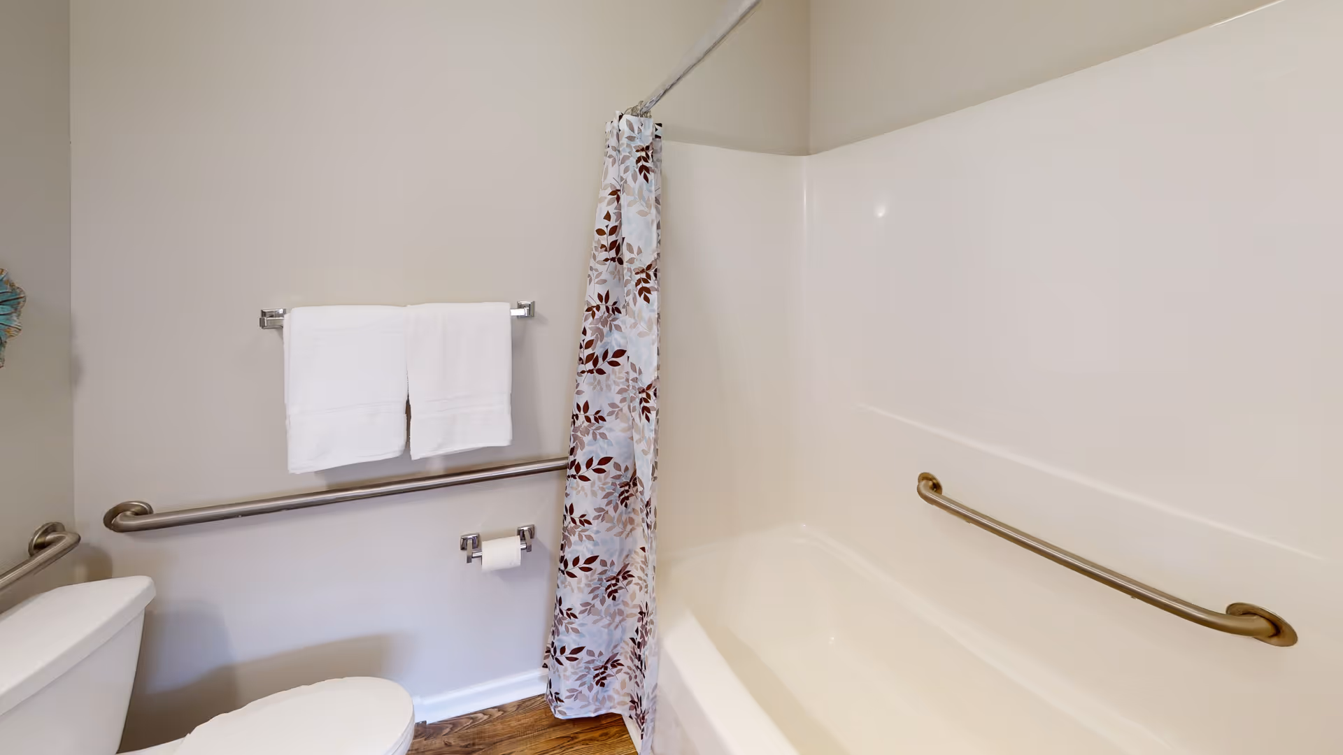 A bathroom featuring a white toilet, a bathtub with a floral patterned shower curtain, white towels hanging on a towel rack, and stainless steel grab bars on the walls. The floor has a wood-like finish.