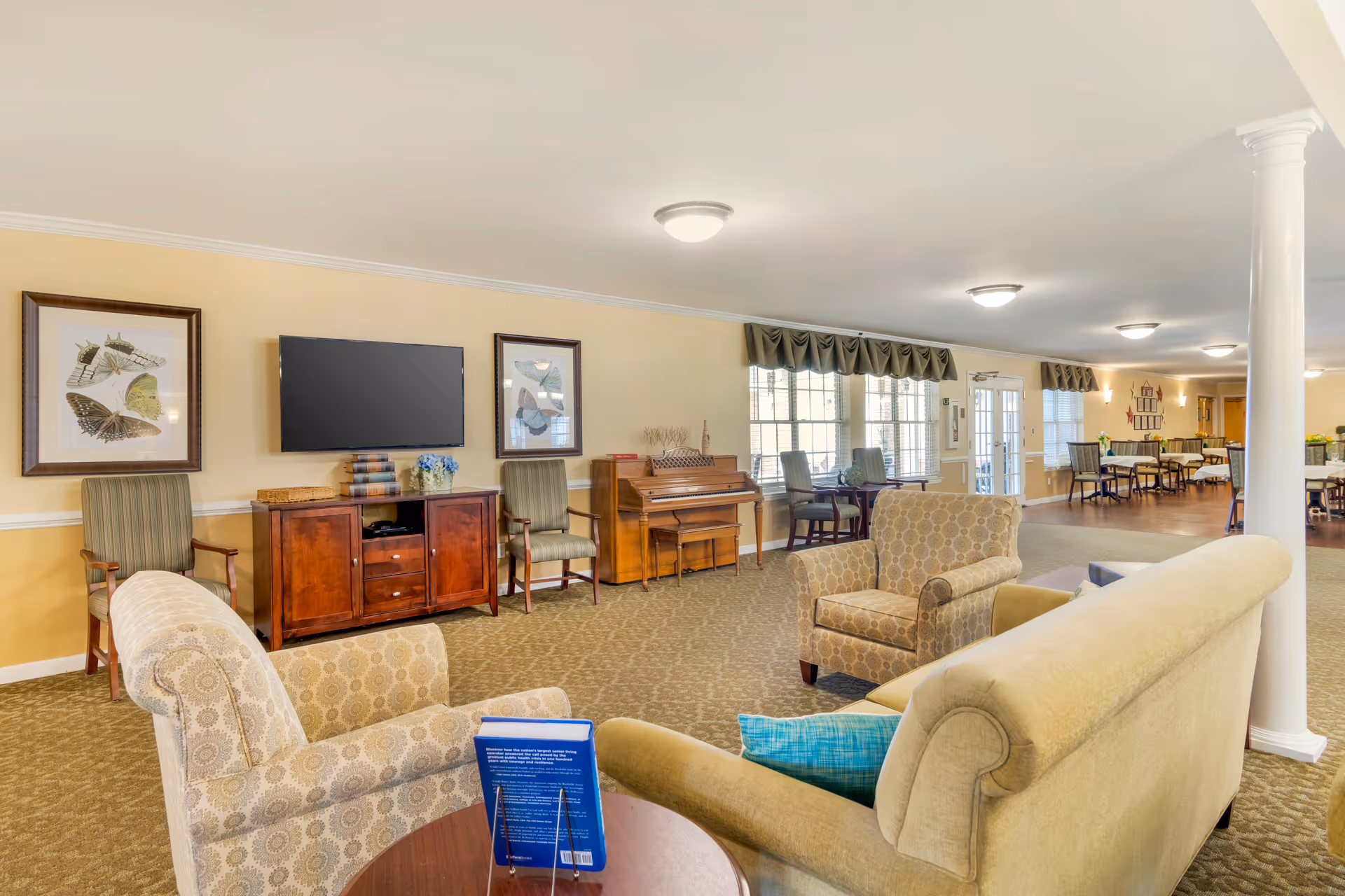 A spacious senior living facility common area with beige walls and carpet. The room features comfortable upholstered armchairs and a sofa arranged around a small round table with a book on it. On the far wall, there is a flat-screen TV mounted between two framed butterfly prints, a wooden cabinet, two chairs, and a piano. Large windows with valances allow natural light to fill the room. In the background, there are several dining tables and chairs near the entrance door.