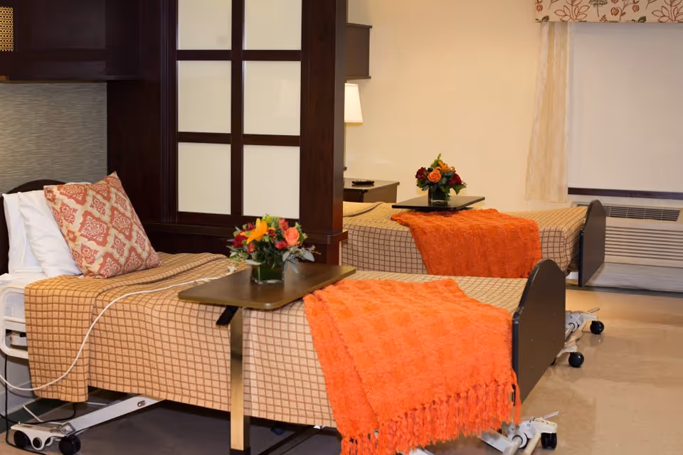 Two hospital-style beds with checkered beige bedspreads and orange throws in a well-lit room. Each bed has a small table with a vase of colorful flowers. There is a decorative wooden partition between the beds and a window with curtains in the background.