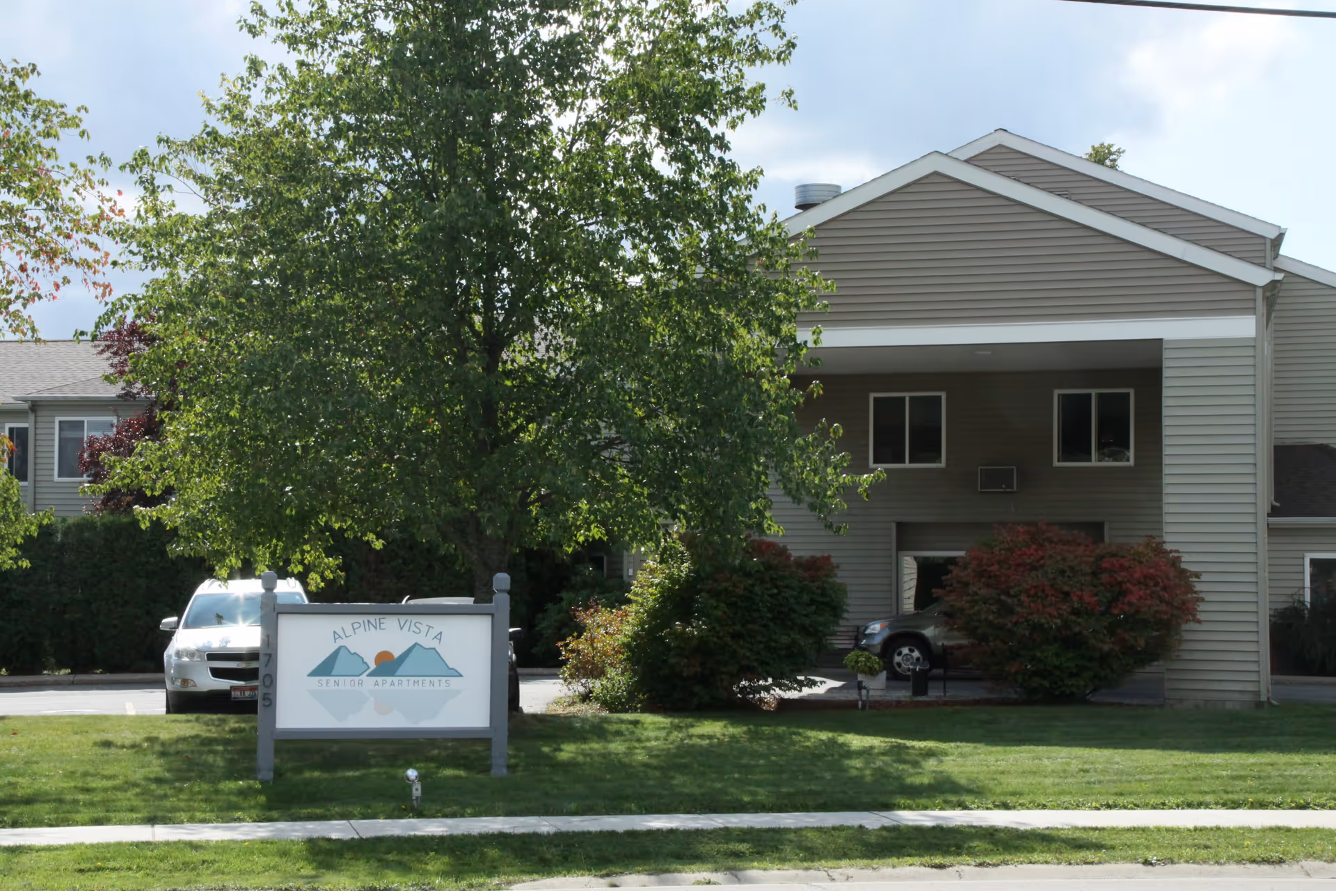 Exterior view of Alpine Vista Senior Apartments building with a large tree and bushes in front, a sign displaying the facility name, and parked cars visible in the background.