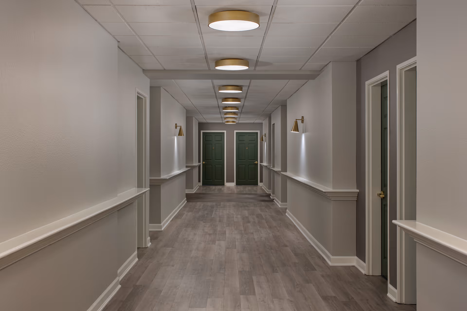 A clean, well-lit hallway in an assisted living facility with light gray walls, wood-look flooring, multiple green doors on both sides, and modern ceiling lights and wall sconces.