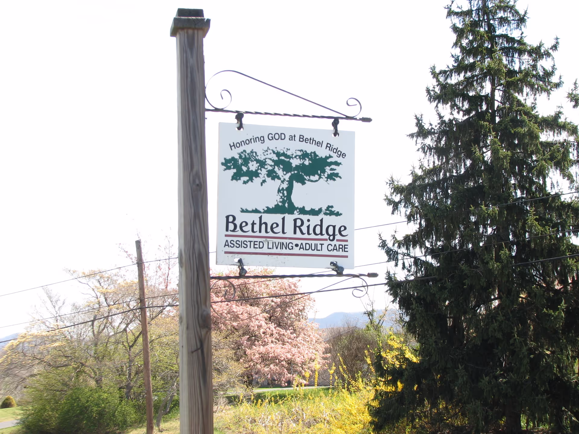 A wooden post with a hanging sign that reads 'Honoring GOD at Bethel Ridge, Bethel Ridge ASSISTED LIVING • ADULT CARE' with a tree graphic above the text. The background shows trees and a clear sky.