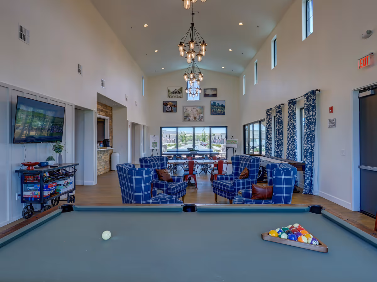 Spacious senior community common room featuring a pool table in the foreground, blue plaid armchairs, dining tables, and large windows.