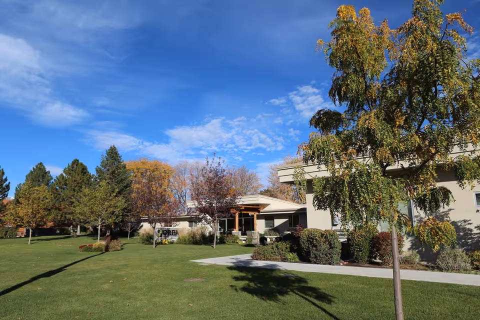 Exterior view of a single-story rehabilitation facility surrounded by green lawn, trees, shrubs, and a walkway under a blue sky.