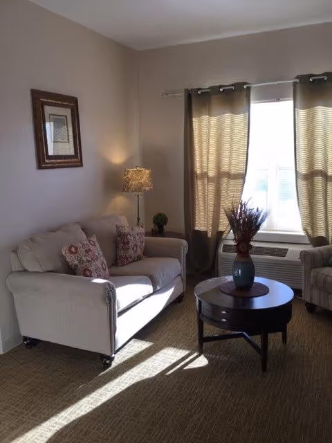A cozy living room with a beige sofa adorned with patterned cushions, a round wooden coffee table with a vase of flowers, a floor lamp, and a window with beige curtains letting in sunlight.