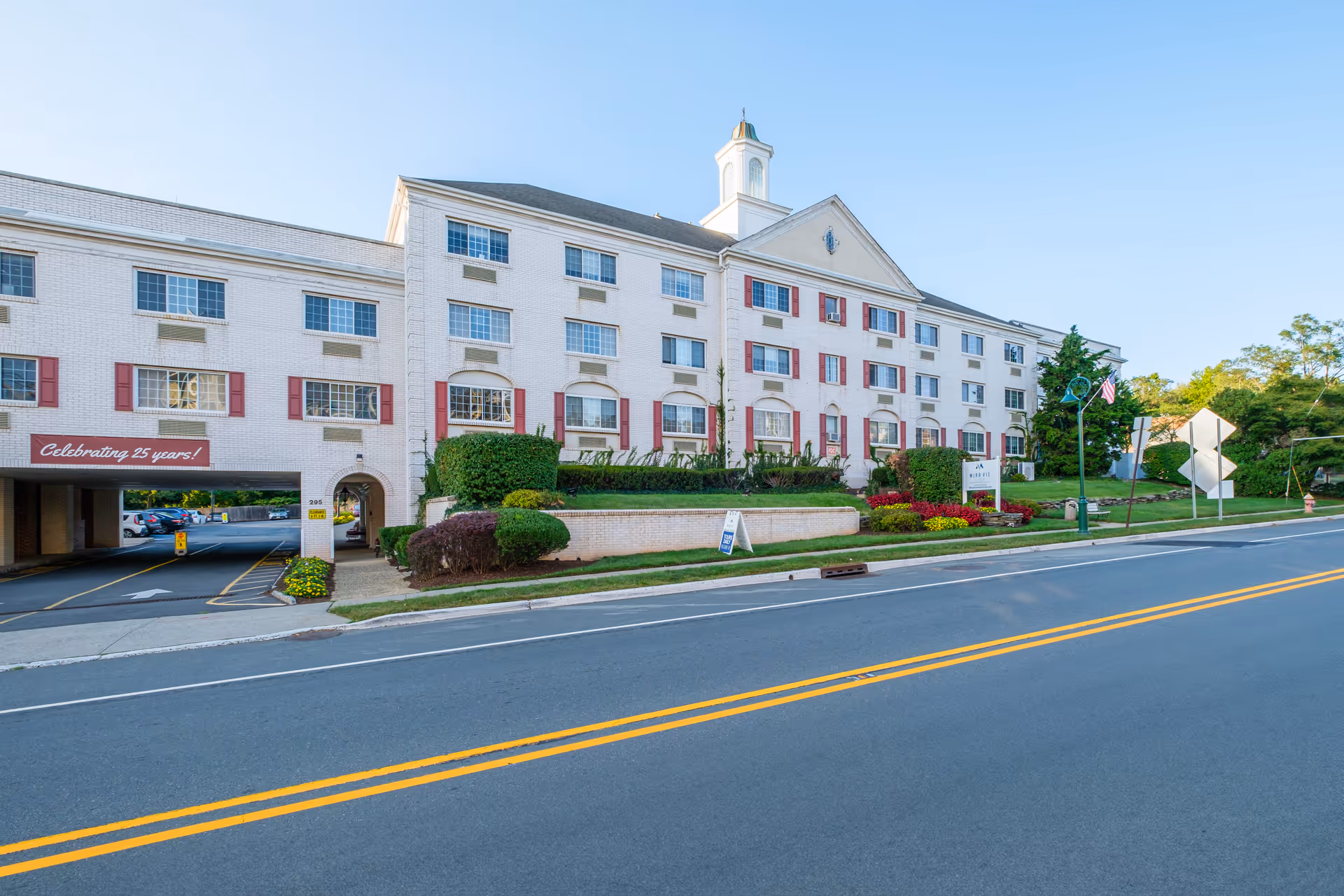 Front exterior of a multi-story white senior living building with red shutters, landscaping, and a street in the foreground.