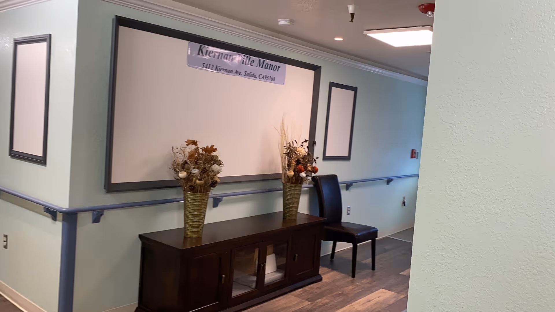 Interior hallway of a senior living facility with light green walls and wooden flooring. A dark wooden cabinet with two vases of dried flowers is placed against the wall, next to a black chair. There are handrails along the walls and framed empty boards hanging above the cabinet.