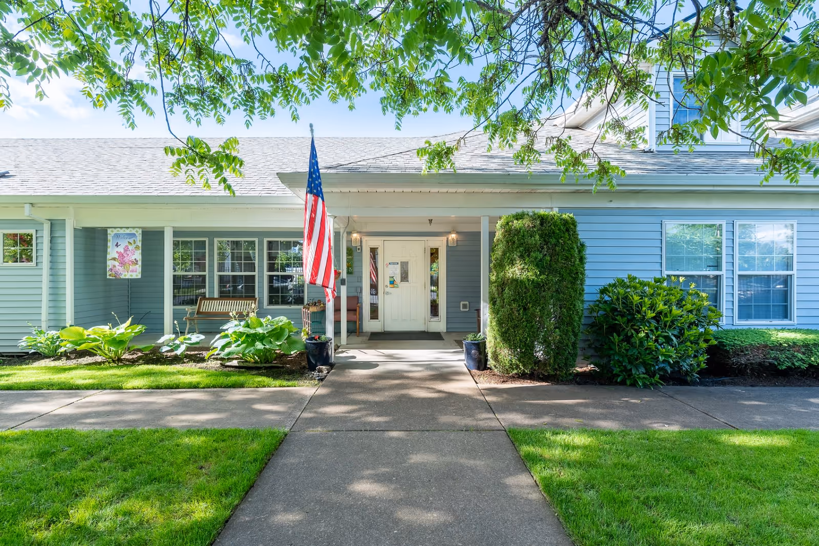 Front exterior view of a single-story building with light blue siding, white trim, and a gray shingled roof. A concrete walkway leads to a covered entrance with double white doors. An American flag is displayed on a pole near the entrance. There are green shrubs and plants along the front of the building, and a wooden bench is visible on the porch. Tree branches with green leaves frame the top of the image.