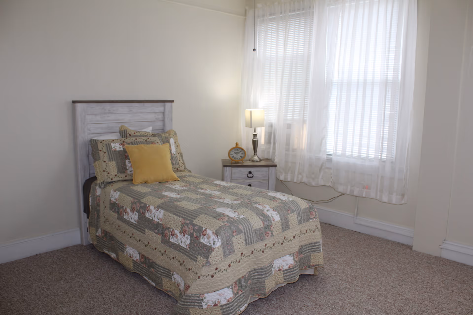 A small bedroom with a single bed covered in a patchwork quilt with floral and striped patterns. The bed has multiple pillows, including a yellow one. Next to the bed is a wooden nightstand with a lamp and a round clock. The room has light-colored walls, carpeted floor, and a window with sheer white curtains allowing natural light to enter.
