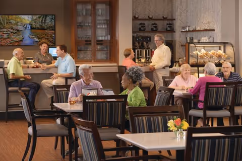 A dining area in a senior living community with several elderly residents sitting at tables and chatting. In the background, a counter with two people talking and a display case with baked goods are visible. The room has warm lighting and a large TV screen showing a nature scene on the wall.