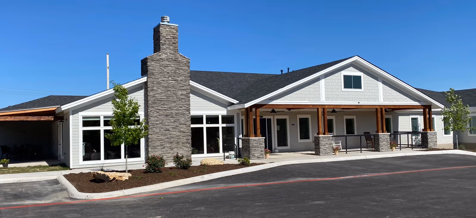 Exterior view of a single-story assisted living facility building with a stone chimney, large windows, a covered porch supported by wooden beams and stone pillars, small trees and shrubs in landscaped beds, and a paved driveway in front under a clear blue sky.