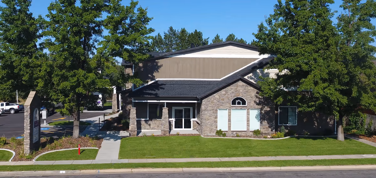 Front exterior view of Layton Park Memory Care facility with a stone and siding facade, surrounded by green trees and a well-maintained lawn, with a sidewalk and parking area visible.