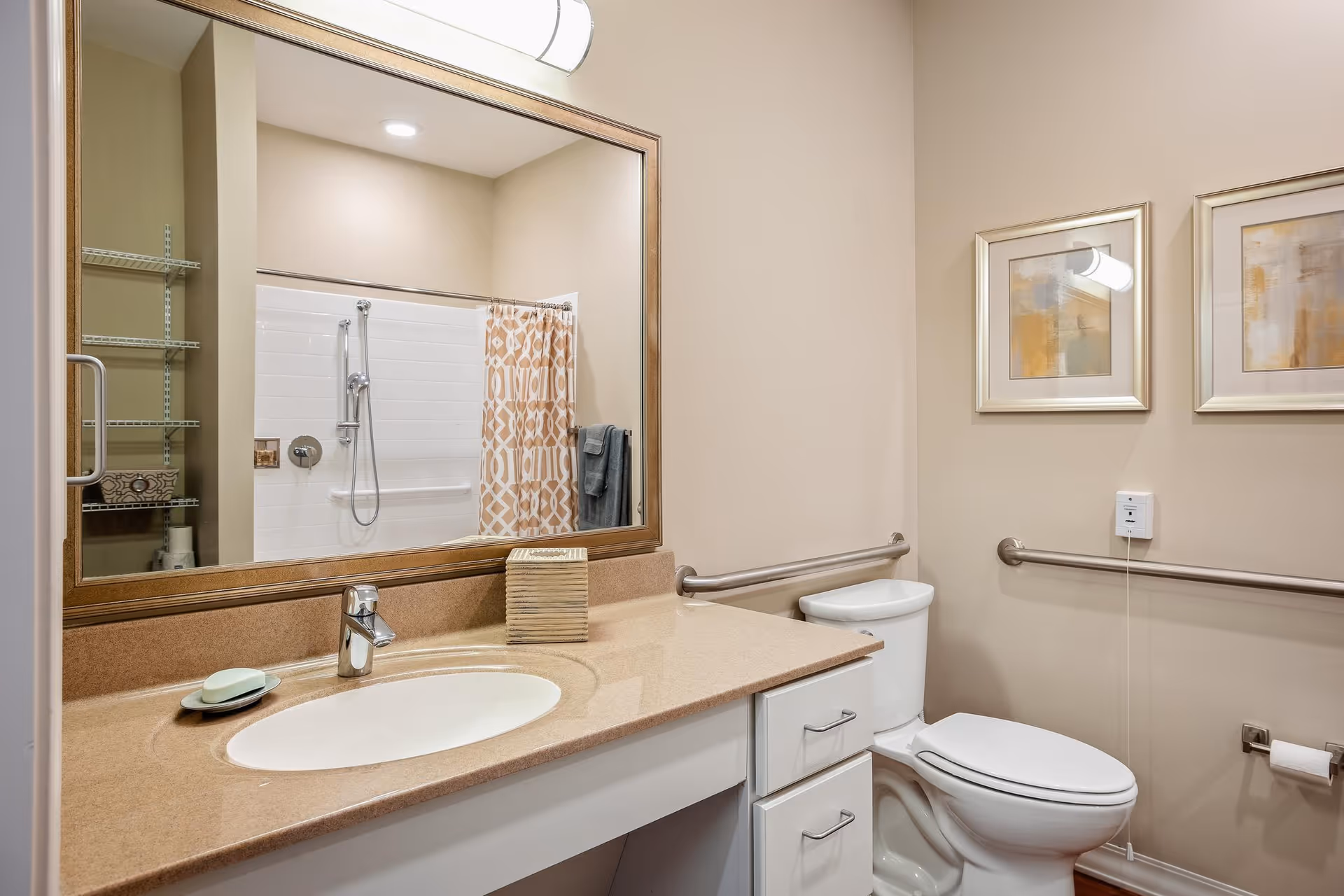 Well-lit accessible bathroom with a sink and vanity beneath a large mirror, a toilet with grab bars, and a shower with a patterned curtain.