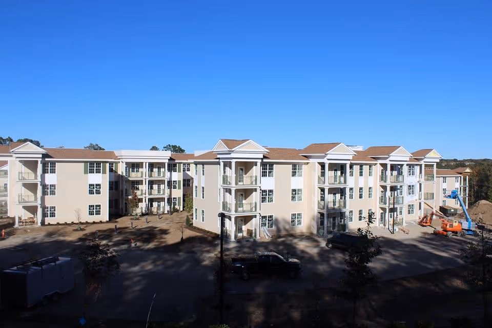 Exterior view of a multi-story senior living facility building under clear blue sky, with balconies, multiple windows, and construction equipment visible on the right side.