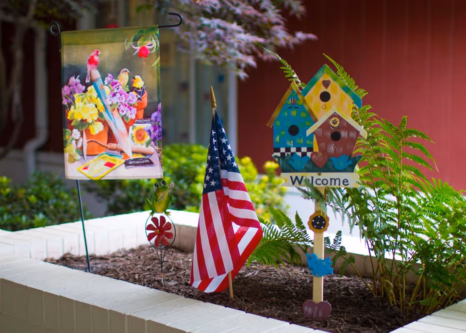 A small garden bed with decorative items including a colorful flag with birds and flowers, a small American flag, a green frog figurine on a red flower, and a wooden birdhouse decoration with a 'Welcome' sign surrounded by green ferns. The background shows a red wall and some greenery.