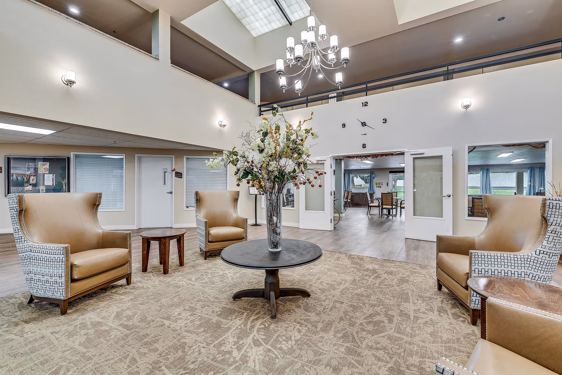 A spacious senior living facility common area with high ceilings and a large chandelier. The room features four tan leather armchairs with patterned sides arranged around a round black table with a tall vase of flowers. There is a beige patterned carpet on the floor and a large wall clock above double doors leading to another room with tables and chairs.
