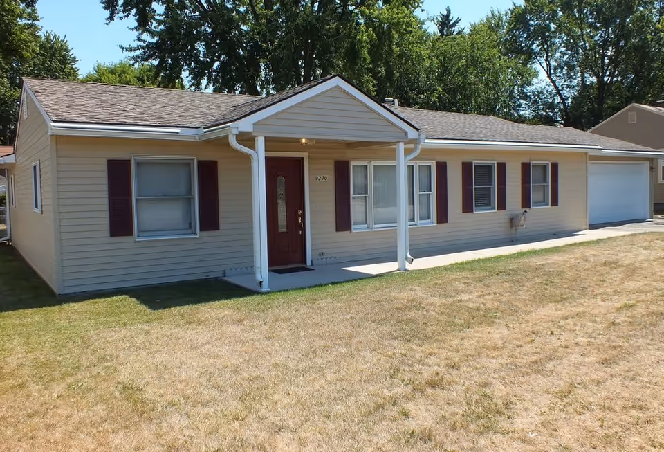 Single-story residential building with beige siding, a brown front door, several windows with maroon shutters, a small covered porch supported by two white columns, and a garage on the right side. The lawn in front is dry and patchy, and there are tall trees in the background under a clear sky.