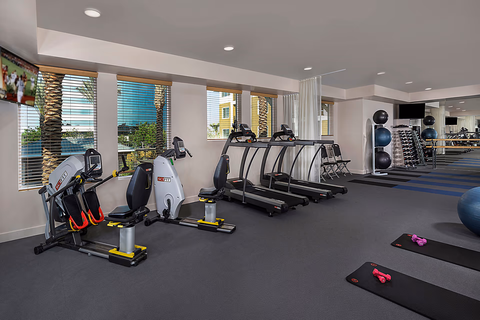 A fitness room with exercise equipment including two treadmills, two recumbent bikes, yoga mats with pink dumbbells, exercise balls, and a rack of dumbbells. Large windows with blinds show palm trees and buildings outside. A wall-mounted TV is visible in the corner.