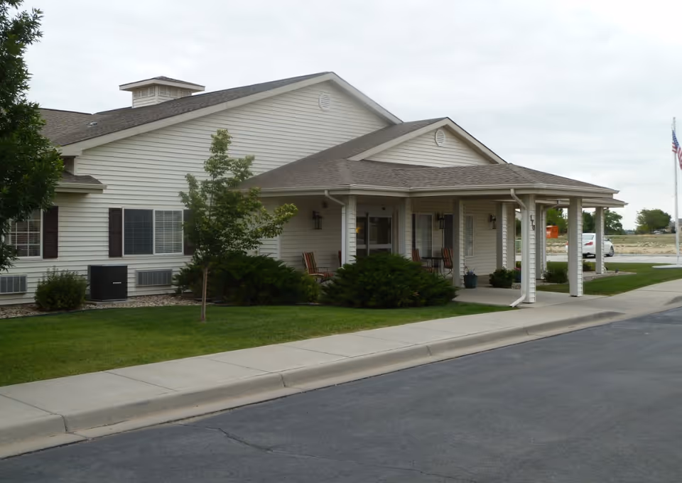 Exterior view of a single-story building with beige siding and a covered entrance porch. There are several chairs on the porch, green bushes, a small tree, and a well-maintained lawn in front. A sidewalk and paved road are visible in the foreground, and an American flag is seen in the background on the right side.