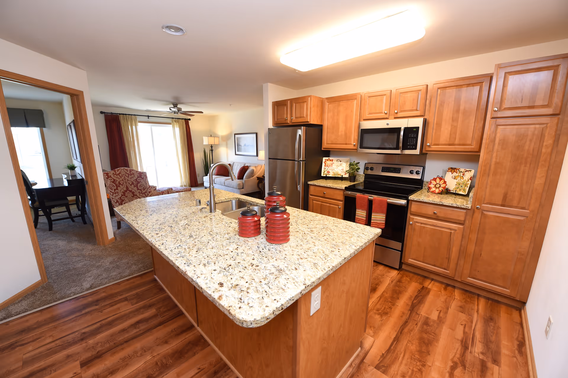 A modern kitchen with wooden cabinets, granite countertops, and stainless steel appliances including a refrigerator, microwave, and stove. The kitchen island has a sink and three red decorative jars. In the background, there is a living room with a sofa, armchair, floor lamp, and large window with curtains. To the left, there is a doorway leading to a carpeted room with a desk and chair.