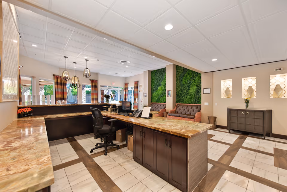 Reception area of Healdsburg Senior Living with a large marble countertop desk, office chair, computer, and phone. The room features a tiled floor with wood accents, pendant lights hanging from the ceiling, and a seating area with two brown tufted sofas against a green plant wall. There are windows with striped curtains and decorative wall niches with lighting on the right side.