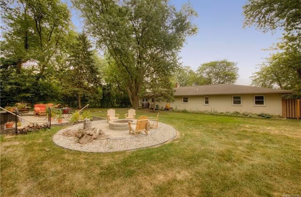 Backyard with a circular fire pit and Adirondack chairs on gravel, a nearby patio with seating, lawn, trees, and a single-story building.