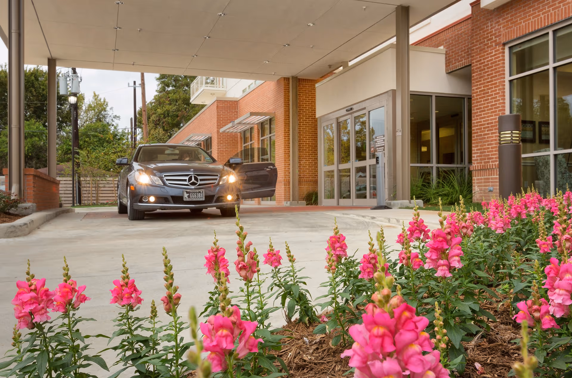 A brick senior living facility entrance with a Mercedes parked under a covered drive and pink flowers in the foreground.