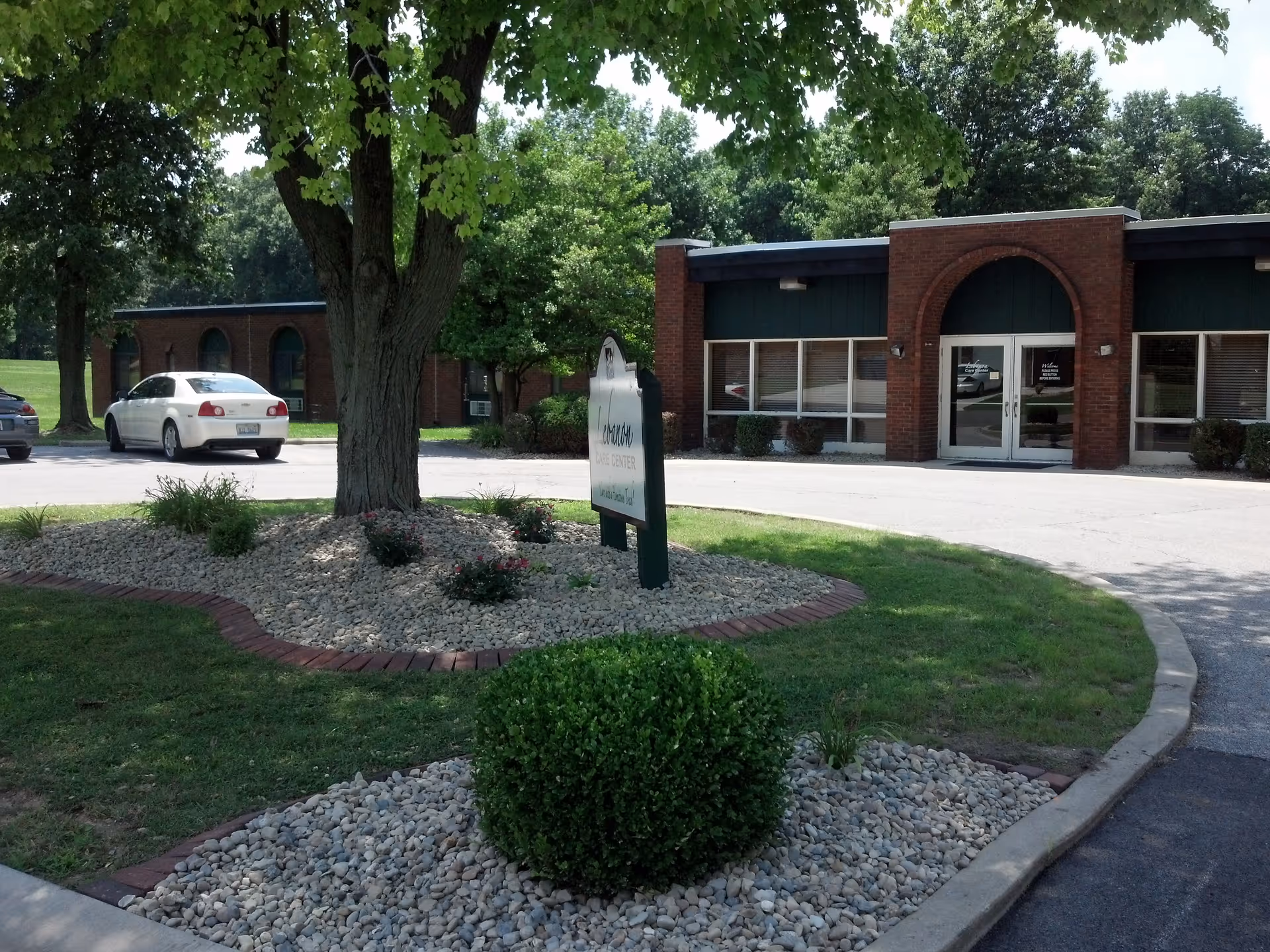 Exterior view of a single-story brick building with large windows and a double glass door entrance. In front of the building is a landscaped area with a tree, bushes, and a sign. There are a few parked cars visible in the parking lot, and trees surround the area.