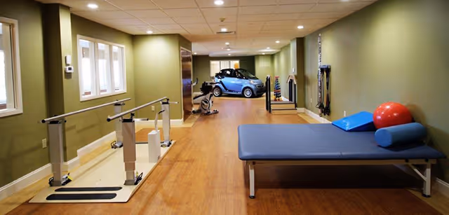 Long rehabilitation/therapy room with parallel bars, a treatment table, exercise equipment and a small mobility car at the far end.