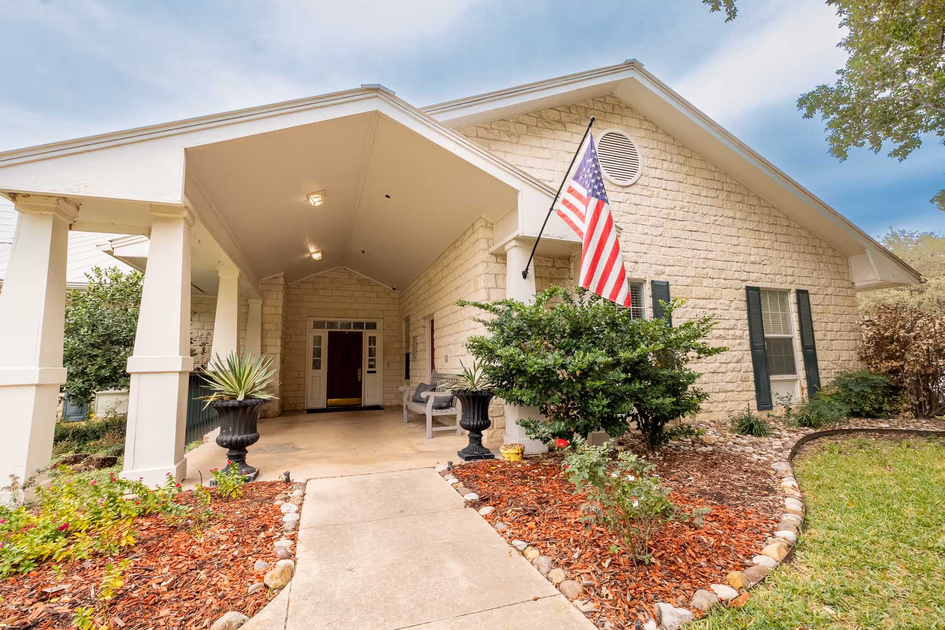 Entrance of a senior living facility with a covered porch supported by white columns, an American flag mounted on the wall, stone exterior walls, a concrete walkway, landscaped garden beds with mulch and plants, and a bench near the door.