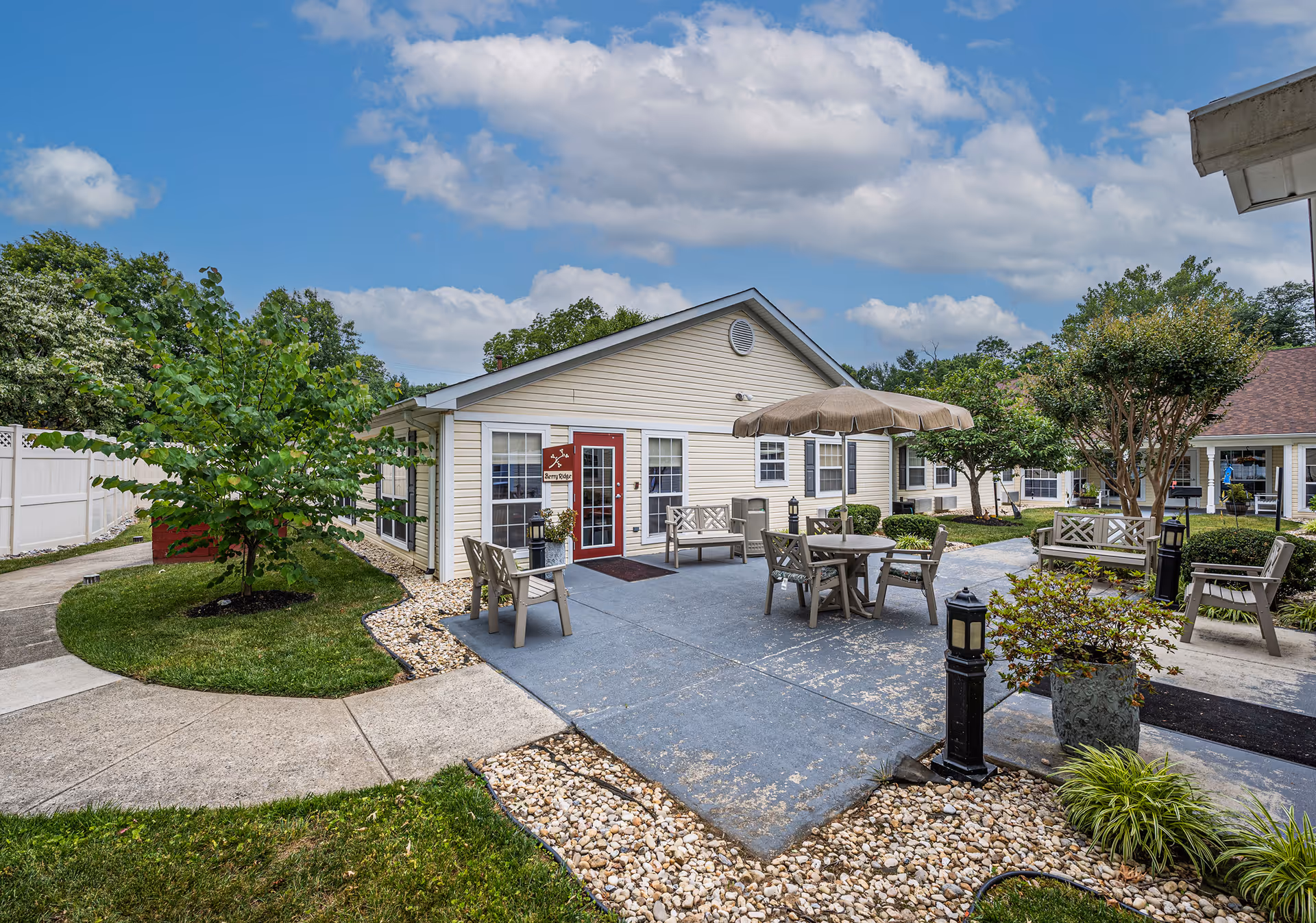Outdoor courtyard and patio area of a single-story memory care building with tables, an umbrella, benches, landscaping and a red entrance door.